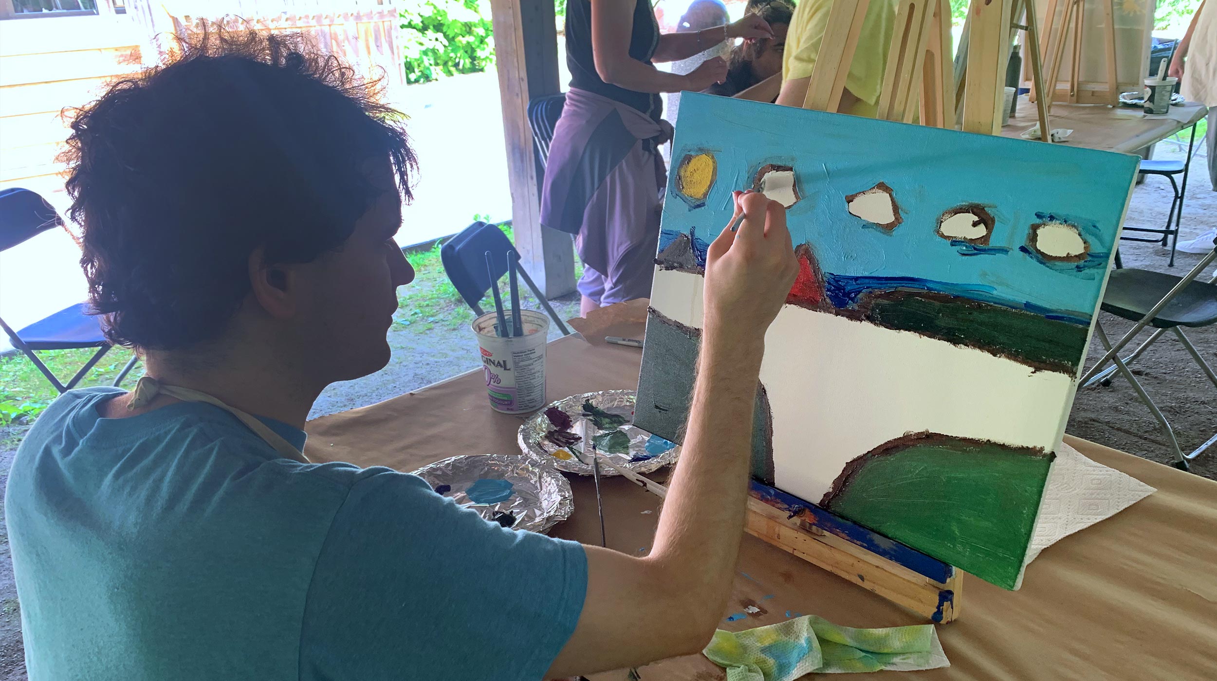 Person painting a landscape scene with blue sky, clouds, and green hills on a canvas outdoors.