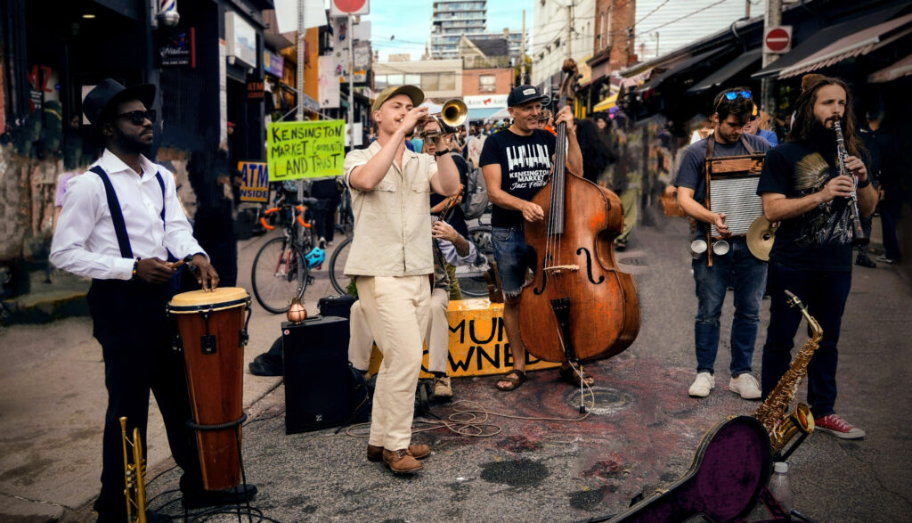 Street musicians performing with trumpet, upright bass, clarinet, washboard, and hand drum in a busy outdoor market street with signs and bicycles in the background.