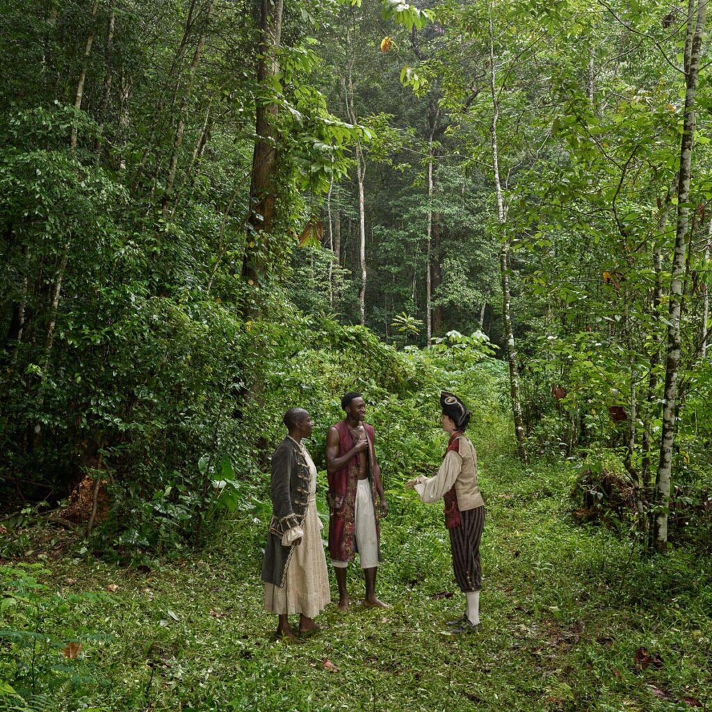 Three figures in historical attire discuss beneath the forest canopy