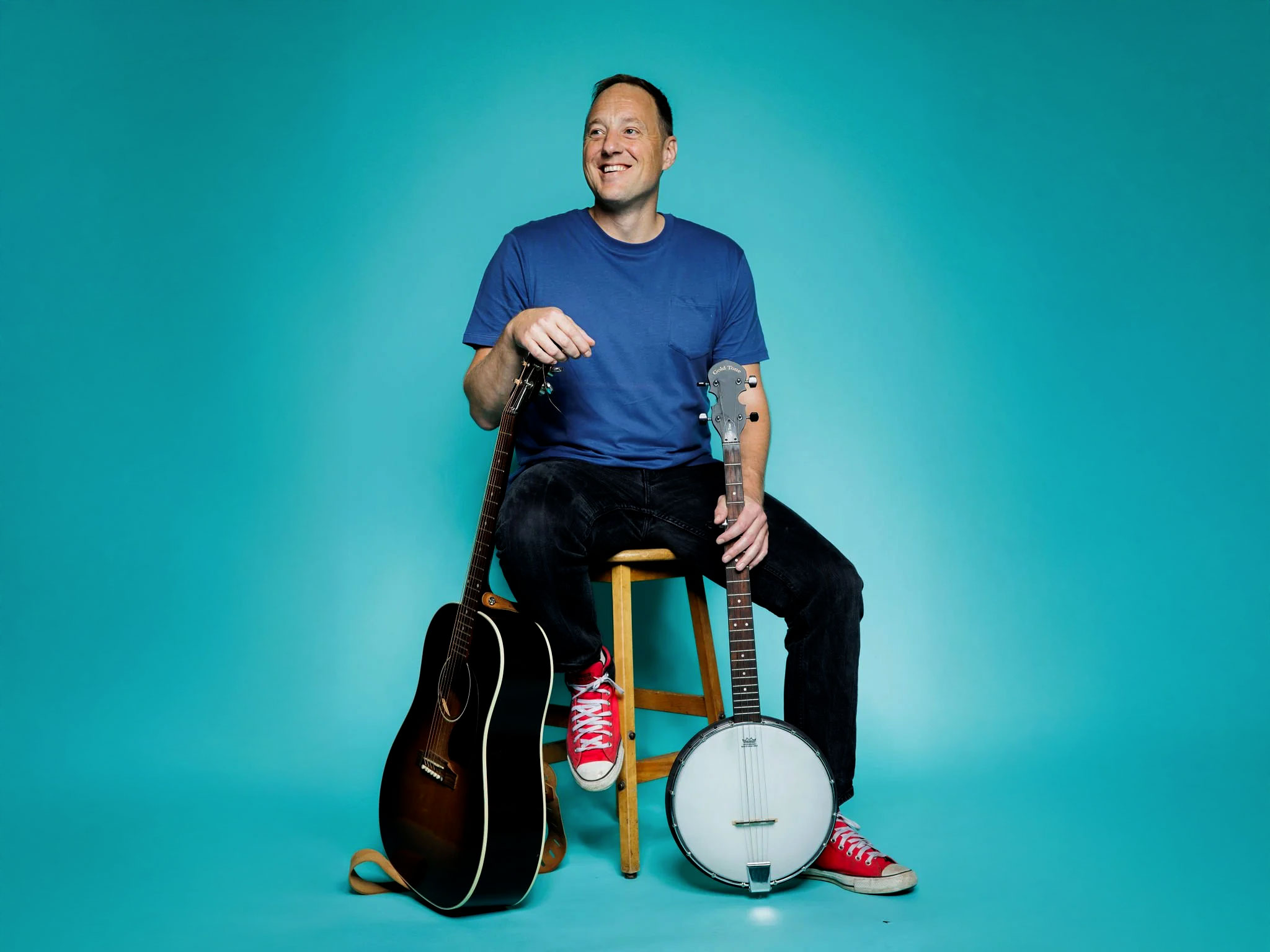 Brendan McLeod sitting on a stool with a guitar on one side and a banjo on the other, against a teal background.