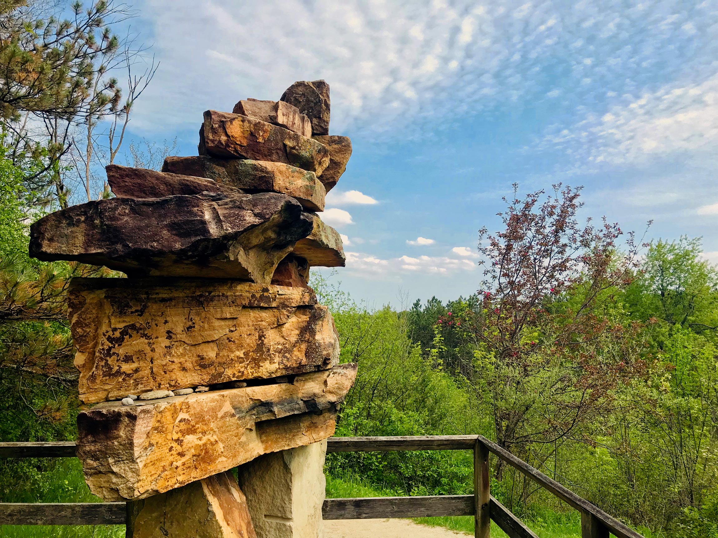 Tall stacked stone sculpture on a wooden platform overlooking green trees under a partly cloudy sky.