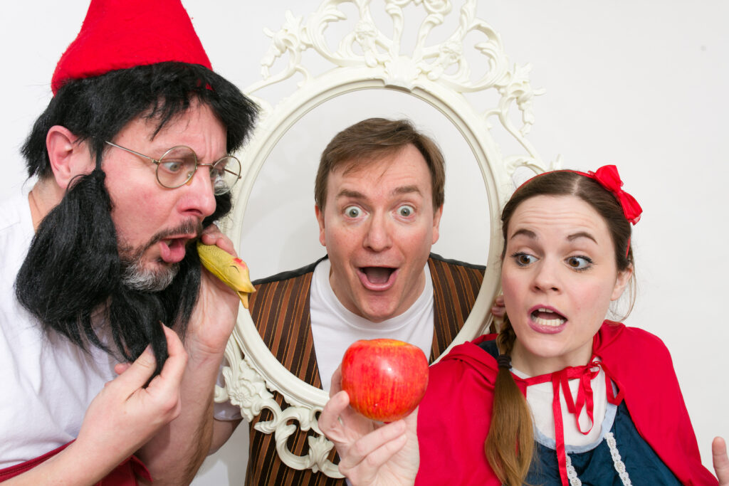 People in fairy-tale themed costumes holding a decorative white frame and a red apple, posing closely together.