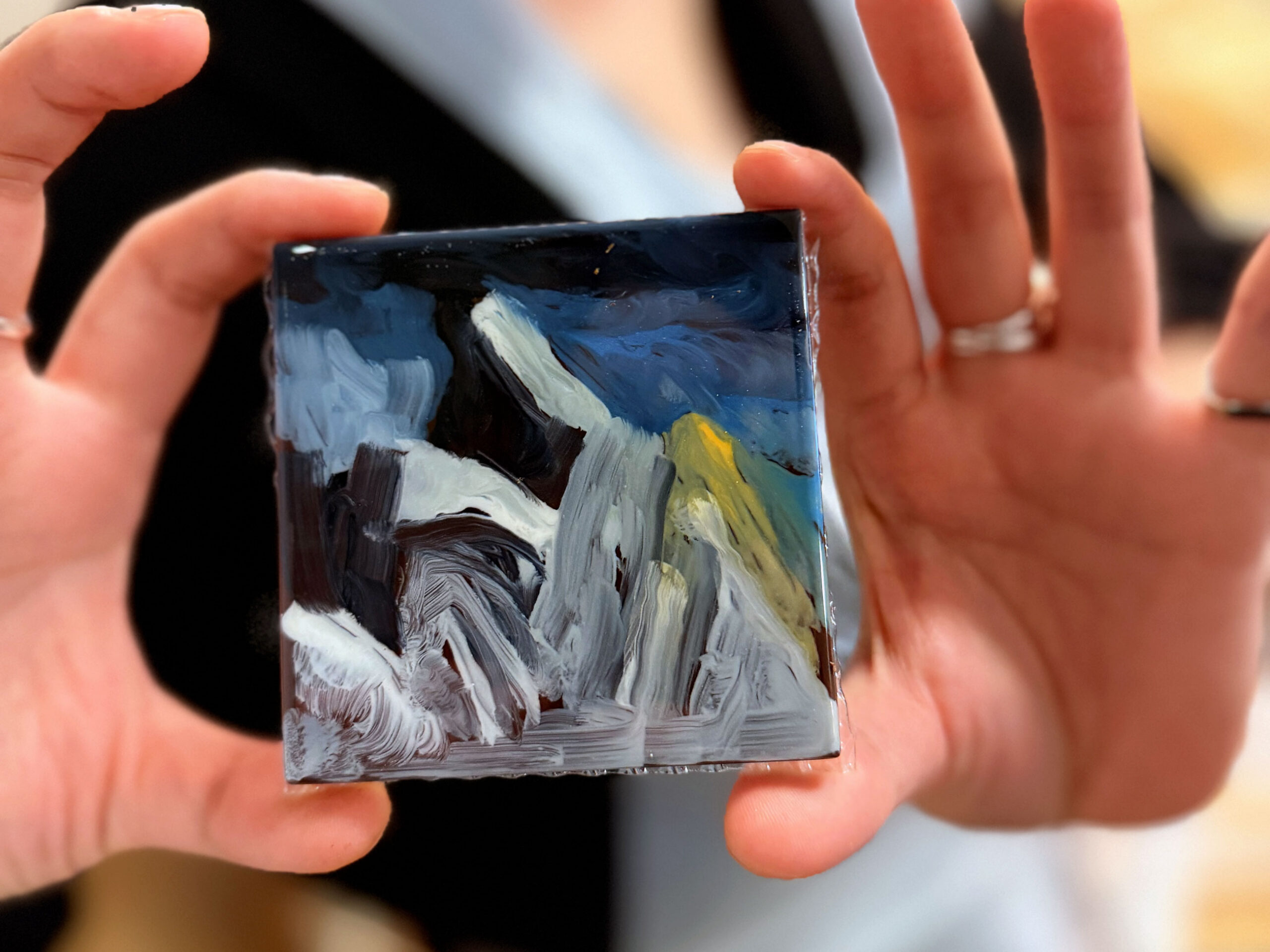 Hands holding a small painted chocolate tile with abstract shapes and colors.