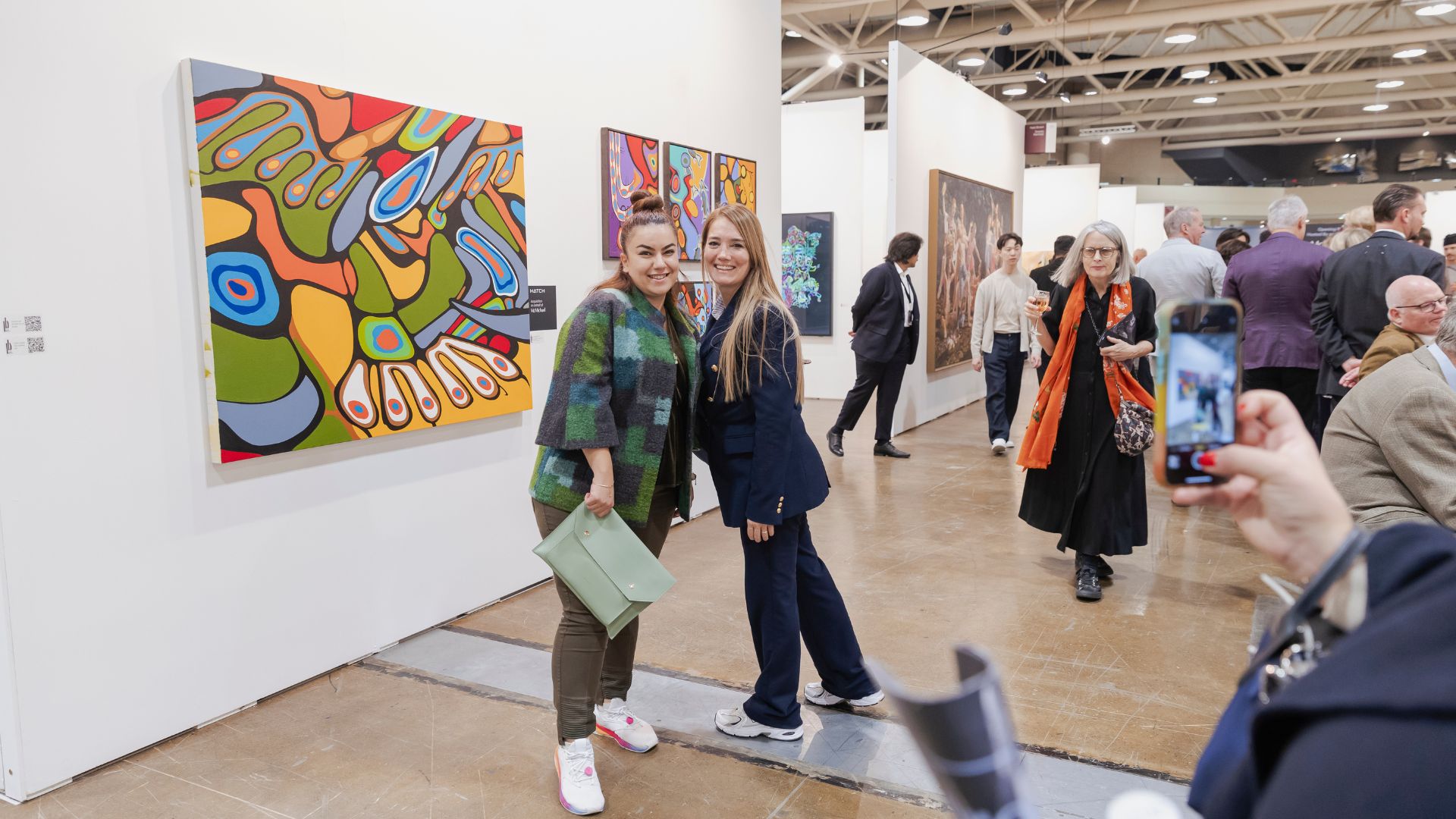 Art gallery hallway with multiple visitors walking past colorful paintings on white walls under bright ceiling lights.