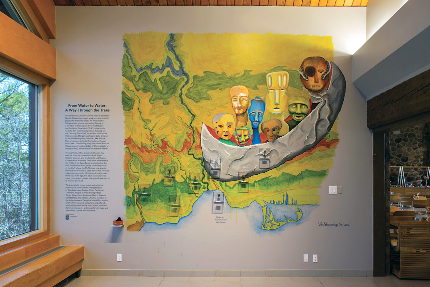 Grande peinture murale colorée présentant des visages stylisés en forme de bateau sur un paysage inspiré d'une carte géographique, exposée sur le mur d'une galerie intérieure ou d'un centre culturel à côté du texte d'exposition.