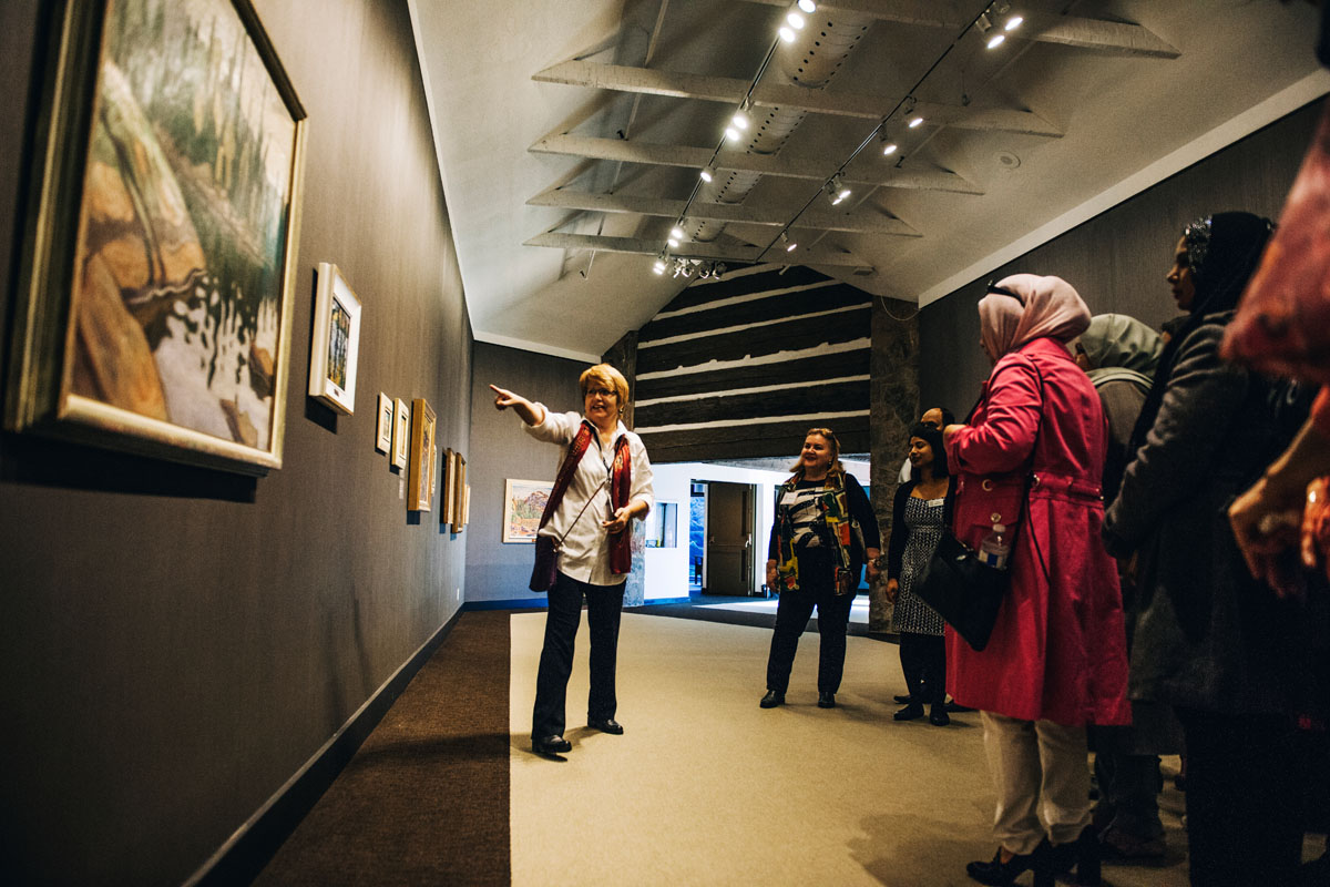 photograph of woman poiting to art in gallery with people gathered around her