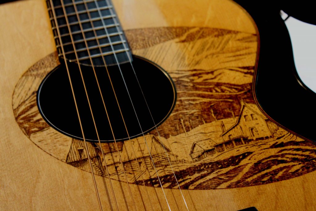 Photograph of the front of a light coloured wood guitar body in close up showing strings and sound hole. The wood around the sound hole shows a scene in sepia tones of primitive buildings with hills in the background.