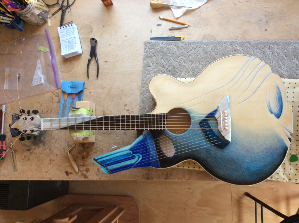 Photograph of a guitar on a work bench seen from above. The guitar is lying on a piece of grey carpet with the headstock to the left. It is decorated in an abstract way in shades of blue, black and cream. There is a secondary partial neck with painted strings coming from the guitar body at a 45 degree angle. There are guitar makers tools on the bench.