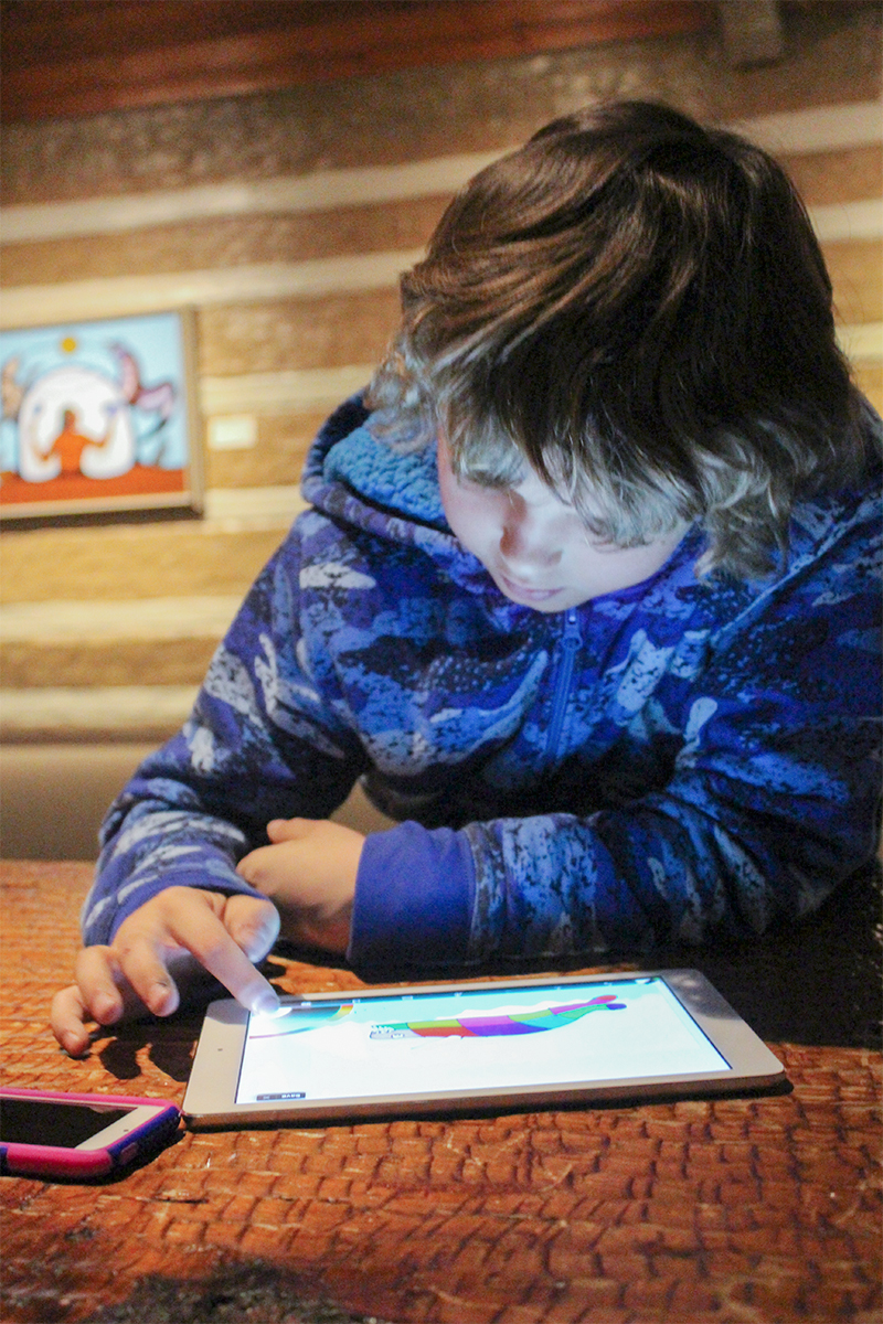 photograph of a child wearing a blue jacket. The child is at a table using an ipad
