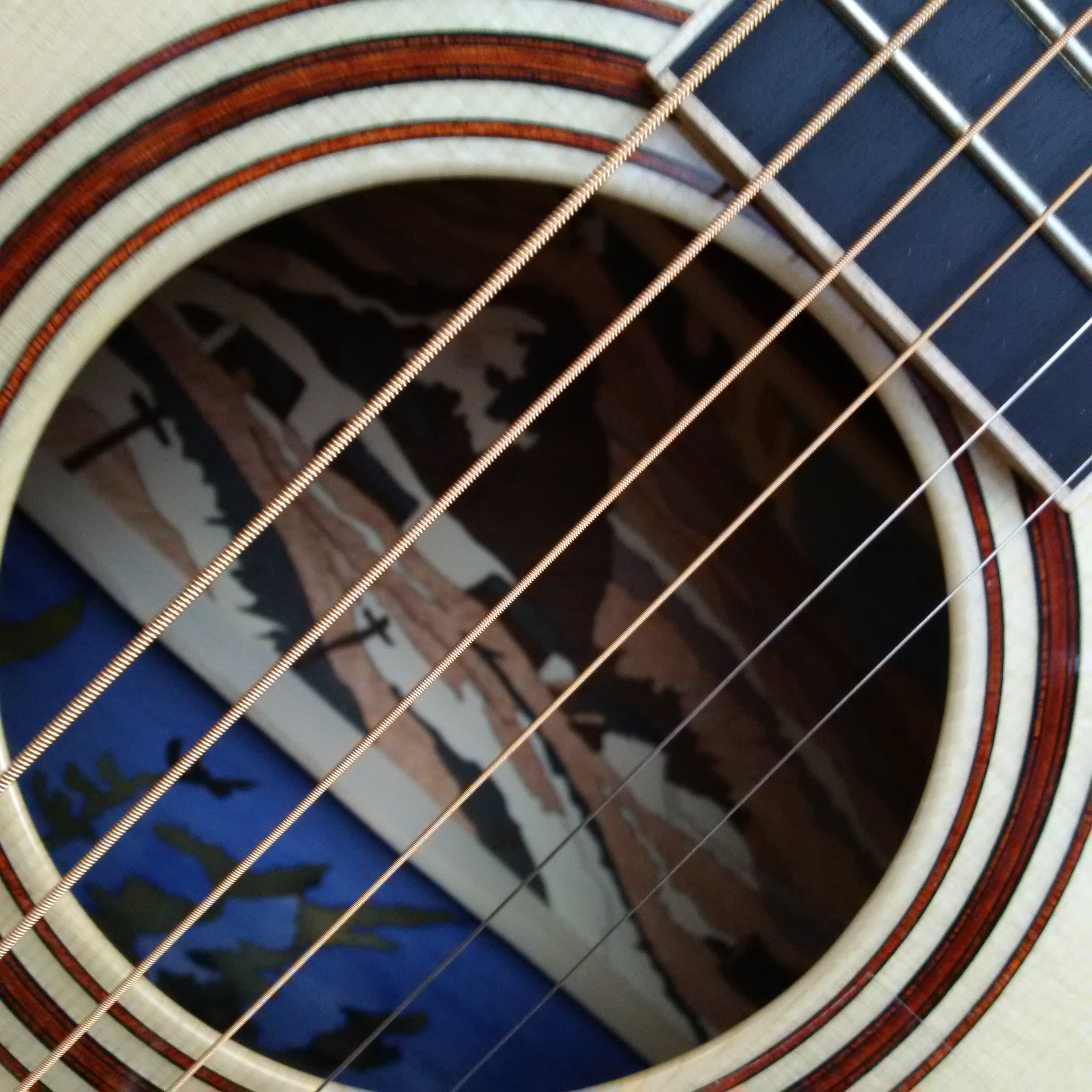 Extreme close up photograph into the interior of a guitar, also showing the sound hole, strings and part of the fretboard. There is a landscape image in the interior in blue, white, black and beige. There are three dark brown circles around the sound hole. The fretboard is dark blue.