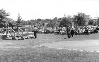 Black and white photograph of a people sitting in chairs on a lawn. There is a platform with people seated in chairs on the left side of the photograph. There are trees in the background.