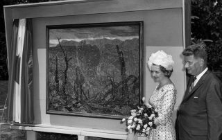 Photograph in black and white of a man and woman standing beside a painting of a landscape in an impressionistic style. The woman is holding a bouquet of roses and wearing a patterned dress and a hat. The man is wearing a suit and tie. There is a curtain pulled aside to the left of the painting.