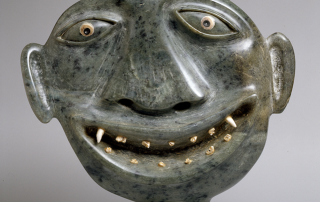 Stone sculpture of a smiling face with tiny legs and protruding ears. There are thirteen teeth in the open mouth.