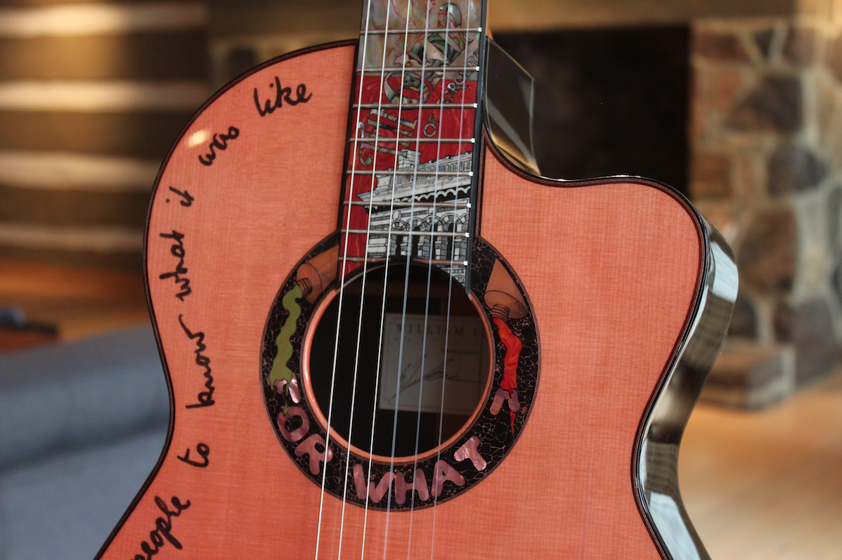 Photograph of mid-section of a guitar. The fretboard is decorated with image of a building. There are words written in black script on the left side of the light orange sounding board and words in capitals and images in a ring around the sound hole.