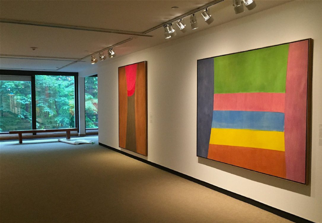 Photograph of two paintings in an abstract style hanging on a wall. The larger painting is colour blocks of pink, orange, yellow, blue, purple and green There is a window to the left with a view of trees.