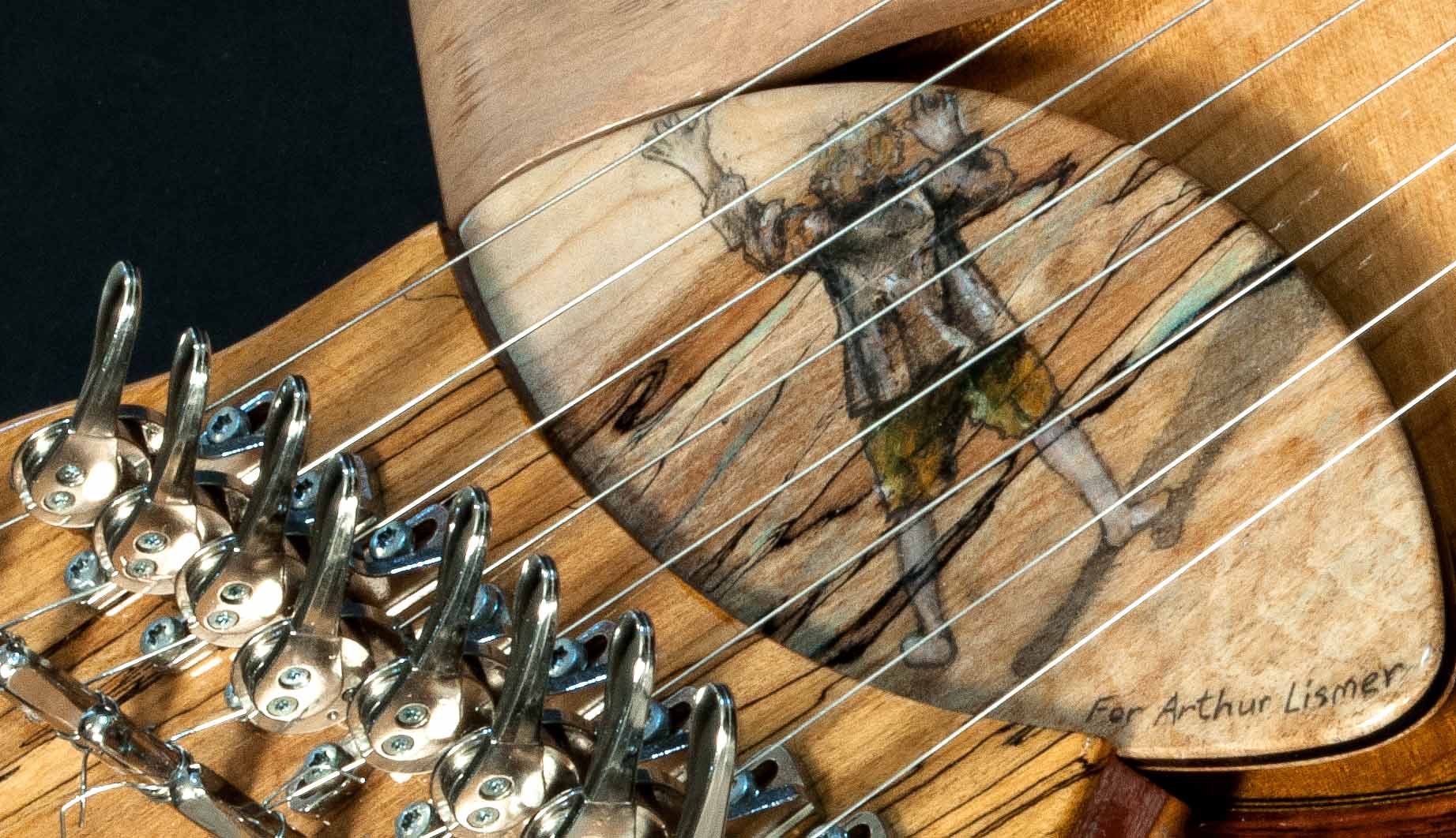 Close-up photograph of head of guitar with strings and metal tuners; detail of wooden insert showing back of man and the words "For Arthur Lismer" at the bottom.